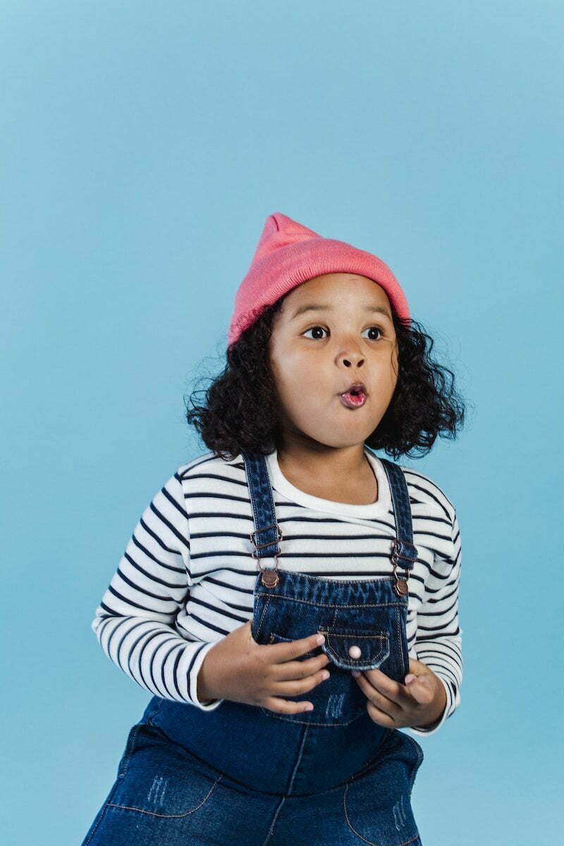 Cute amazed African American girl in cool outfit and pink hat staring away with astonishment while standing on blue background