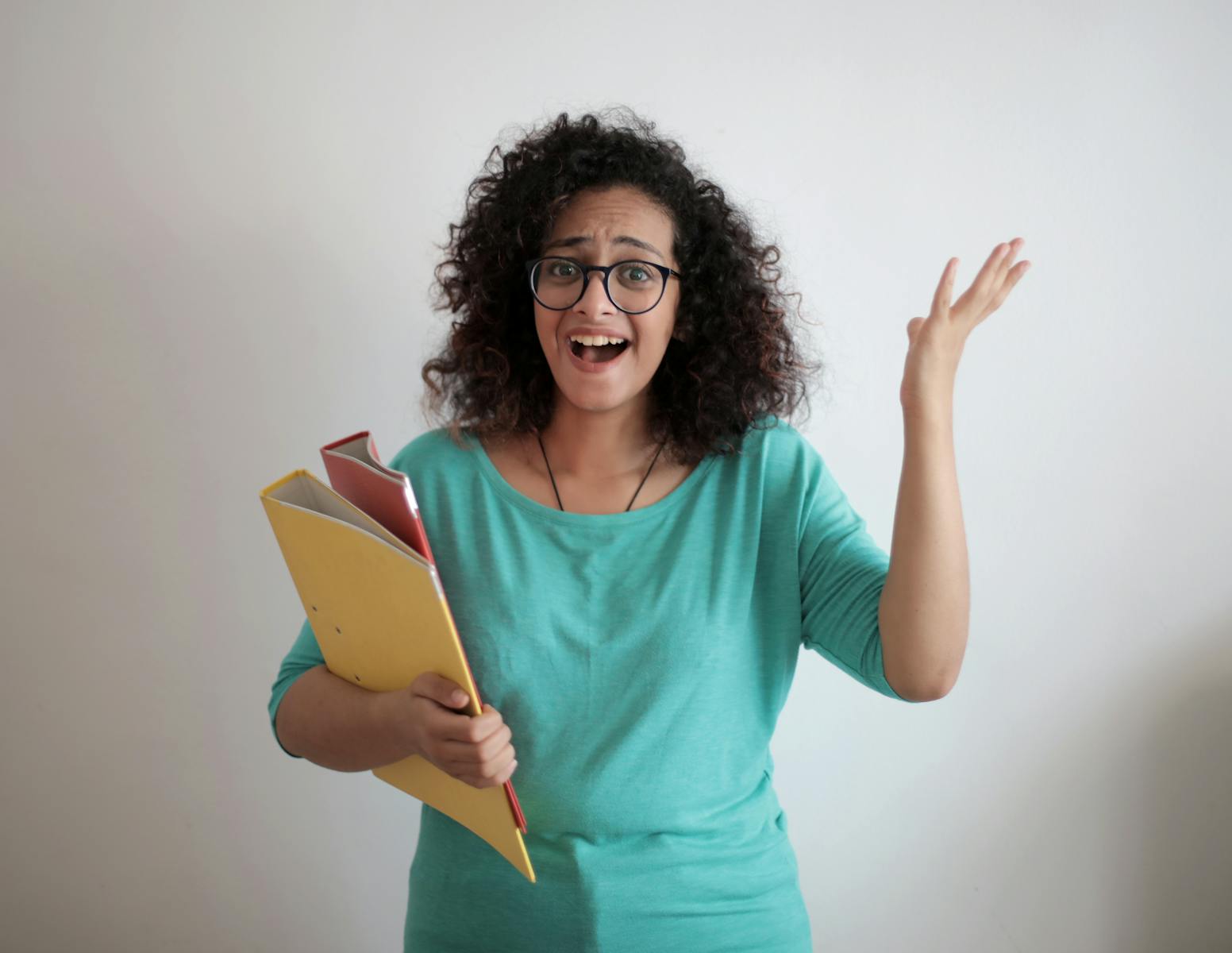 Dissatisfied annoyed woman with mouth opened wearing glasses and turquoise blouse looking away and screaming while standing against white wall with folders of documents and having problems in work